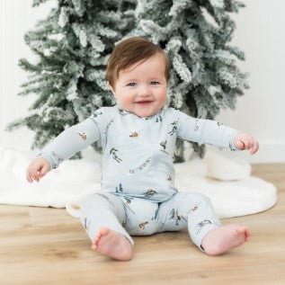 Baby in a light blue onesie with animal prints sitting on a wooden floor in front of a decorated Christmas tree.