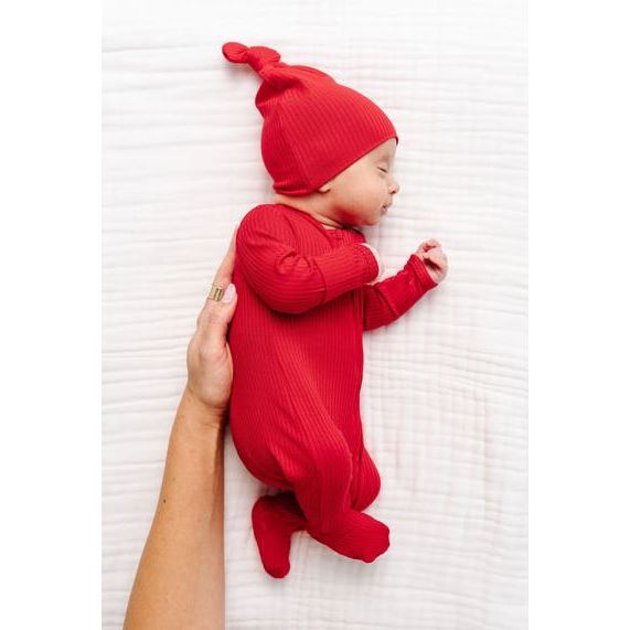 A newborn baby wearing a matching red ribbed knit hat and footed sleeper, lying on a white textured blanket while being held by an adult&
