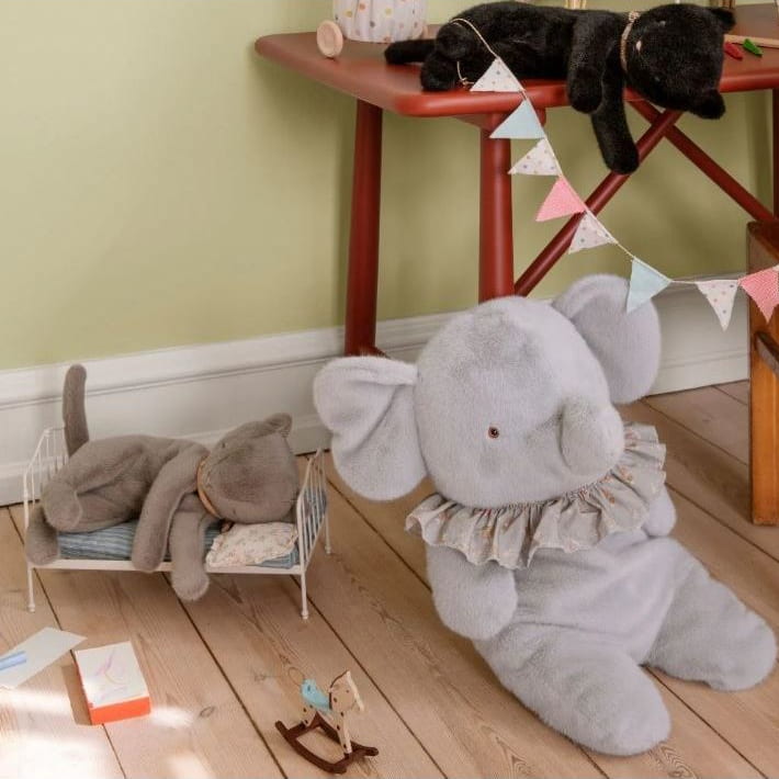 Gray elephant plush toy on a wooden floor with a child's play area in the background.