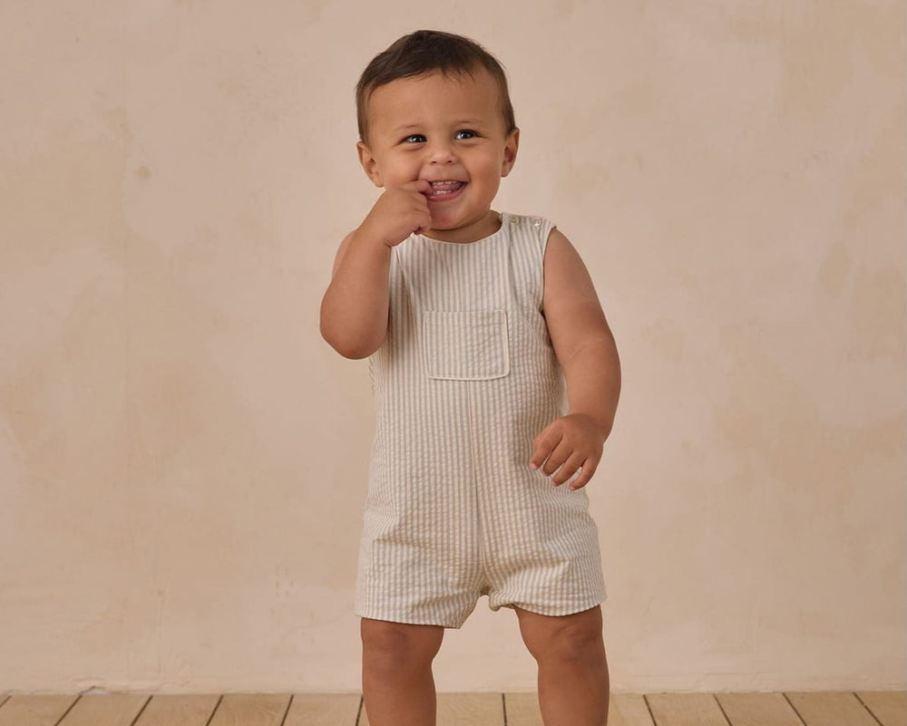 Child wearing a white striped romper standing on a wooden floor with a beige wall background