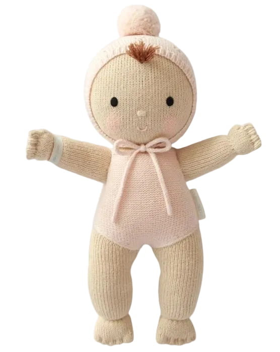 A knitted baby doll toy in soft cream and pale pink, featuring a hood with a pom-pom, black embroidered eyes and mouth, and raised arms, shown against a plain white background.
