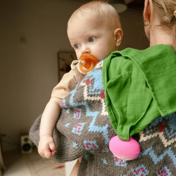 Baby wearing a patterned dress with a pacifier, held by an adult in a green shirt.