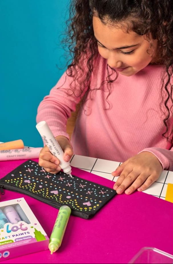 A child in a pink long-sleeve shirt is painting on a black canvas with a white paint tube. The scene is set on a bright magenta surface with a blue background, featuring other paint tubes and a box labeled 'lot of love craft paint'.