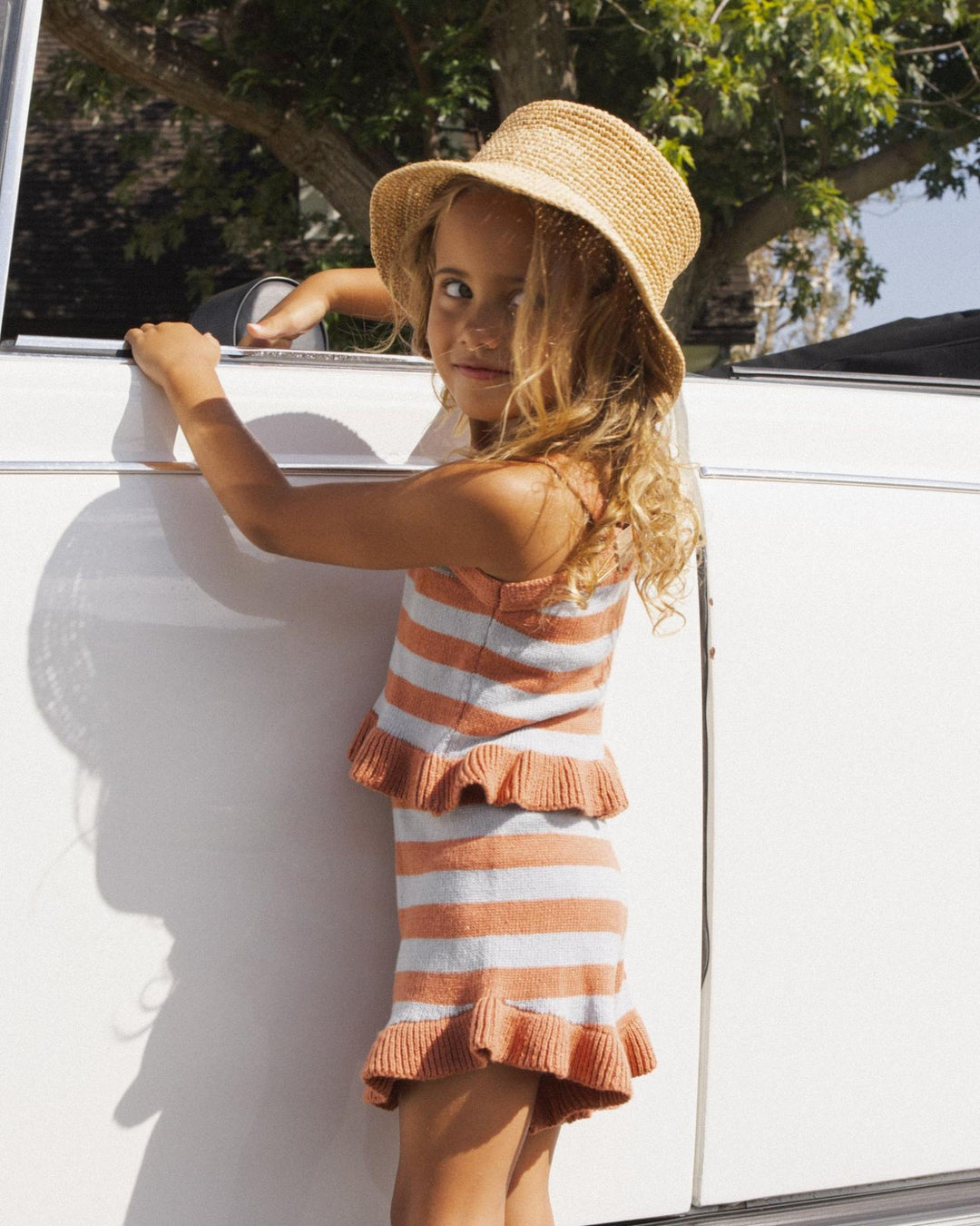Young girl in a striped dress and sun hat leaning against a white vehicle outdoors.