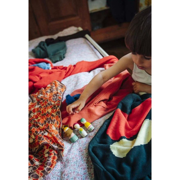 A child sitting on a bed surrounded by colorful clothing and small pill bottles on a patterned sheet in a bedroom setting.