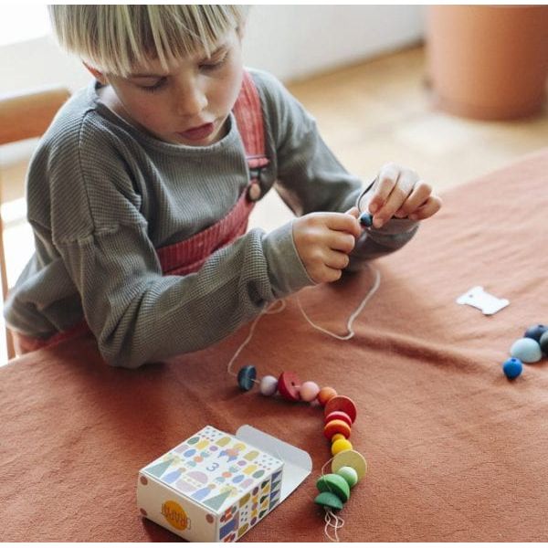 A young child wearing a gray ribbed sweater and red overalls sits at a table, playing with a colorful wooden bead garland and a small patterned box.