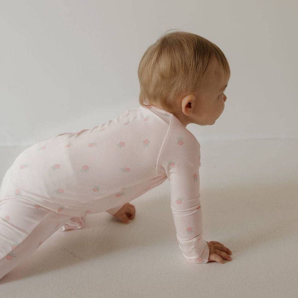 A baby crawling on a light neutral floor wearing a pale pink long-sleeve one-piece with small pink polka dots, photographed in a minimal studio setting.