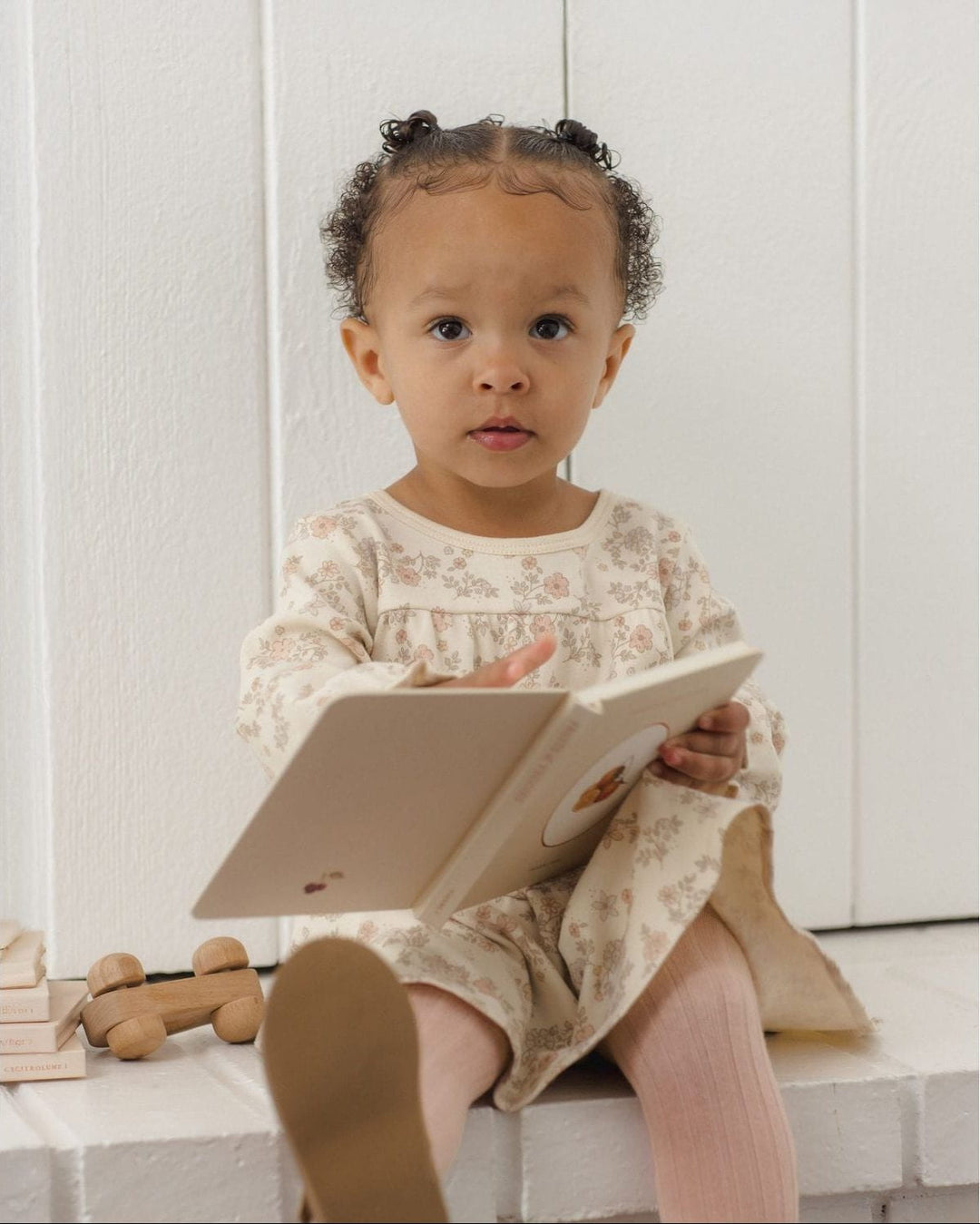 A toddler girl with curly hair sits on a white brick ledge reading a book. She is wearing a cream-colored long-sleeve dress with a delicate pink and green floral pattern, pink tights, and brown shoes.