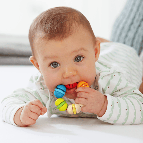 A baby lying on their stomach, chewing on a colorful wooden bead teether toy. The baby is wearing a light green and white striped long-sleeve romper against a soft, bright, neutral background.