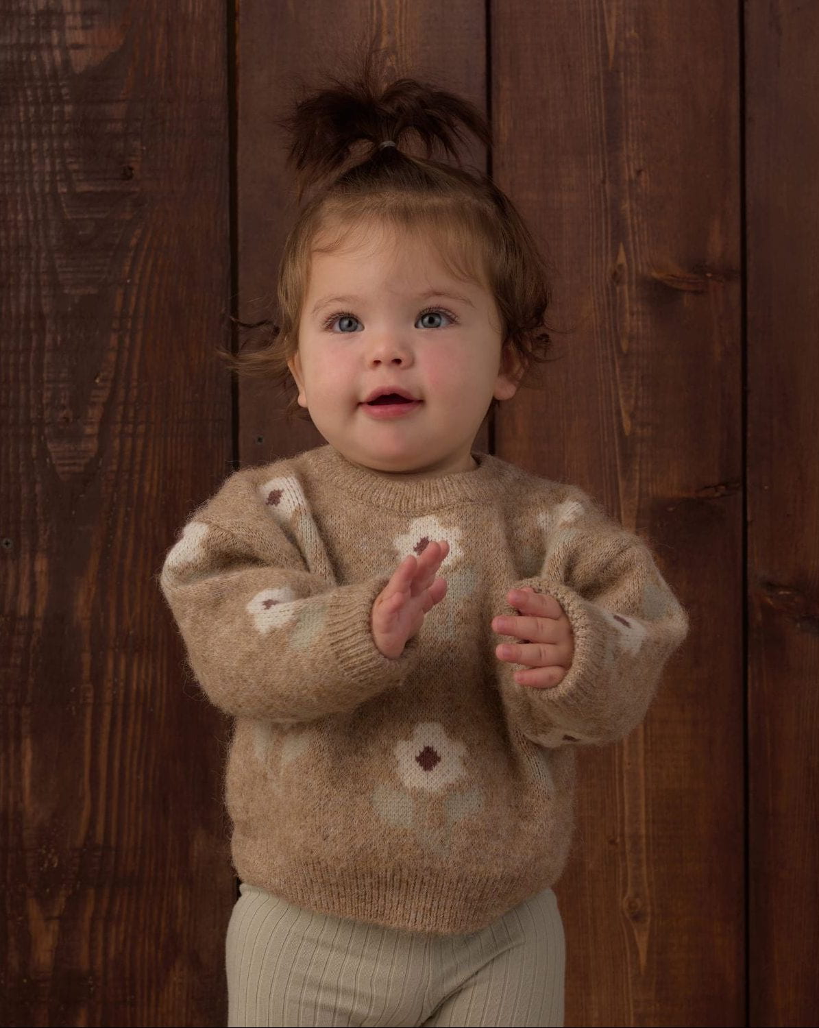 A toddler girl wearing a fuzzy beige knit sweater with white floral patterns, standing in front of a dark brown wooden plank background.