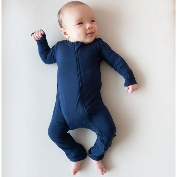 A baby lying on their back wearing a navy blue long-sleeve zip-up footed sleeper, photographed against a plain off-white background.