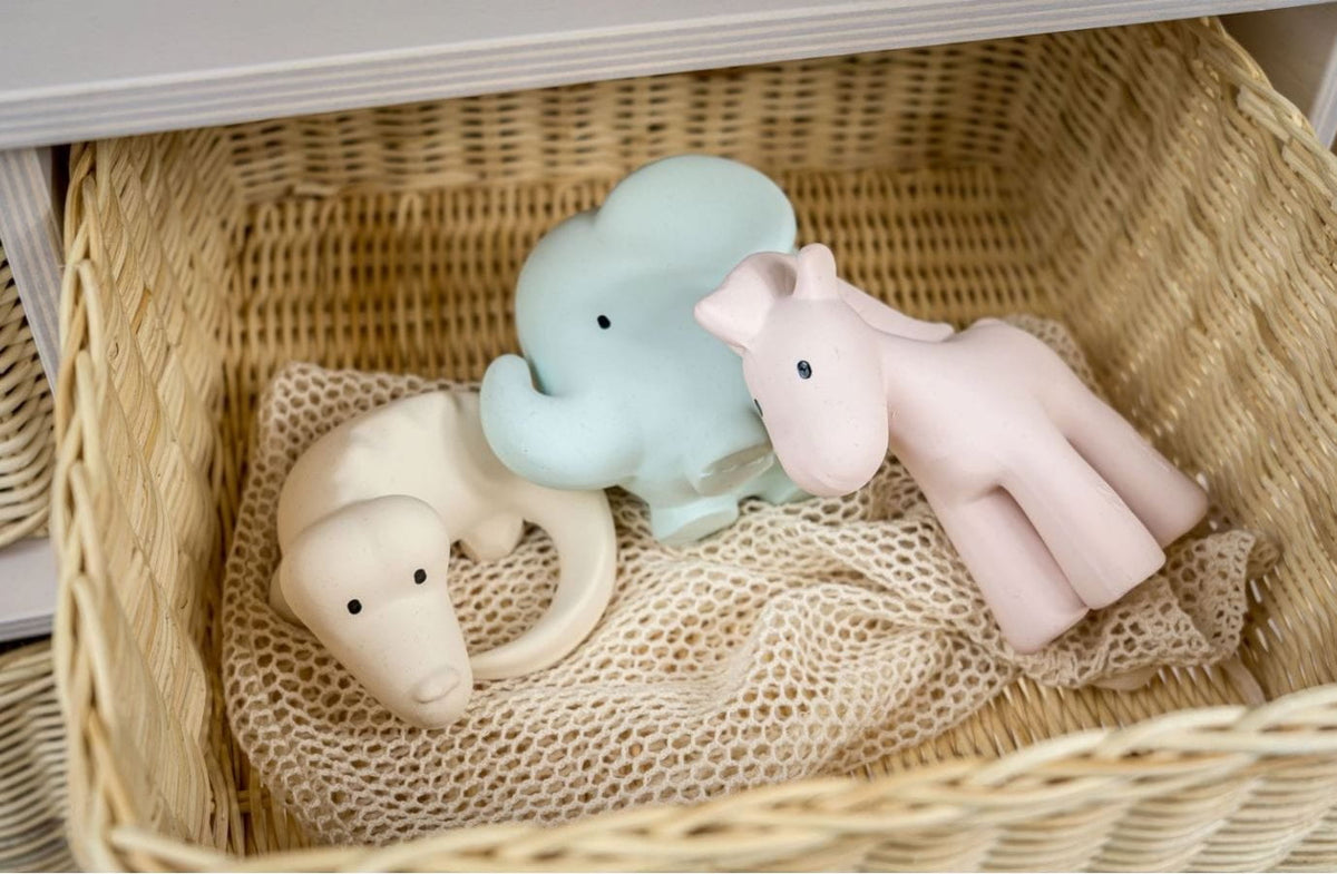 Three soft animal-shaped baby teething toys resting in a woven wicker basket on a mesh cloth. The toys include a pale blue elephant, a cream elephant, and a pale pink giraffe.