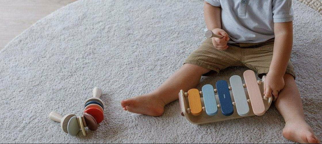 A toddler sits on a round rug playing with a wooden xylophone toy, with a matching wooden toy rattle nearby on a light wood floor.