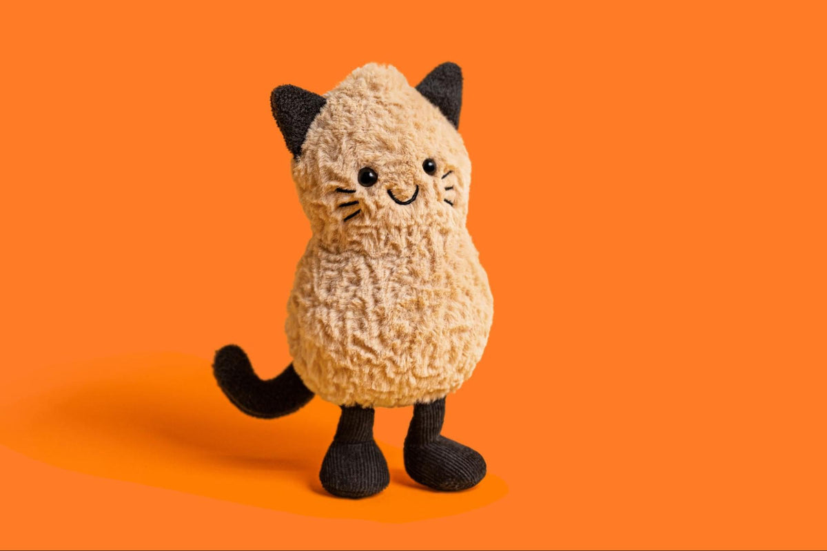 A plush cat toy with a textured cream-colored body, black ears, paws, and tail, standing against a solid orange background.