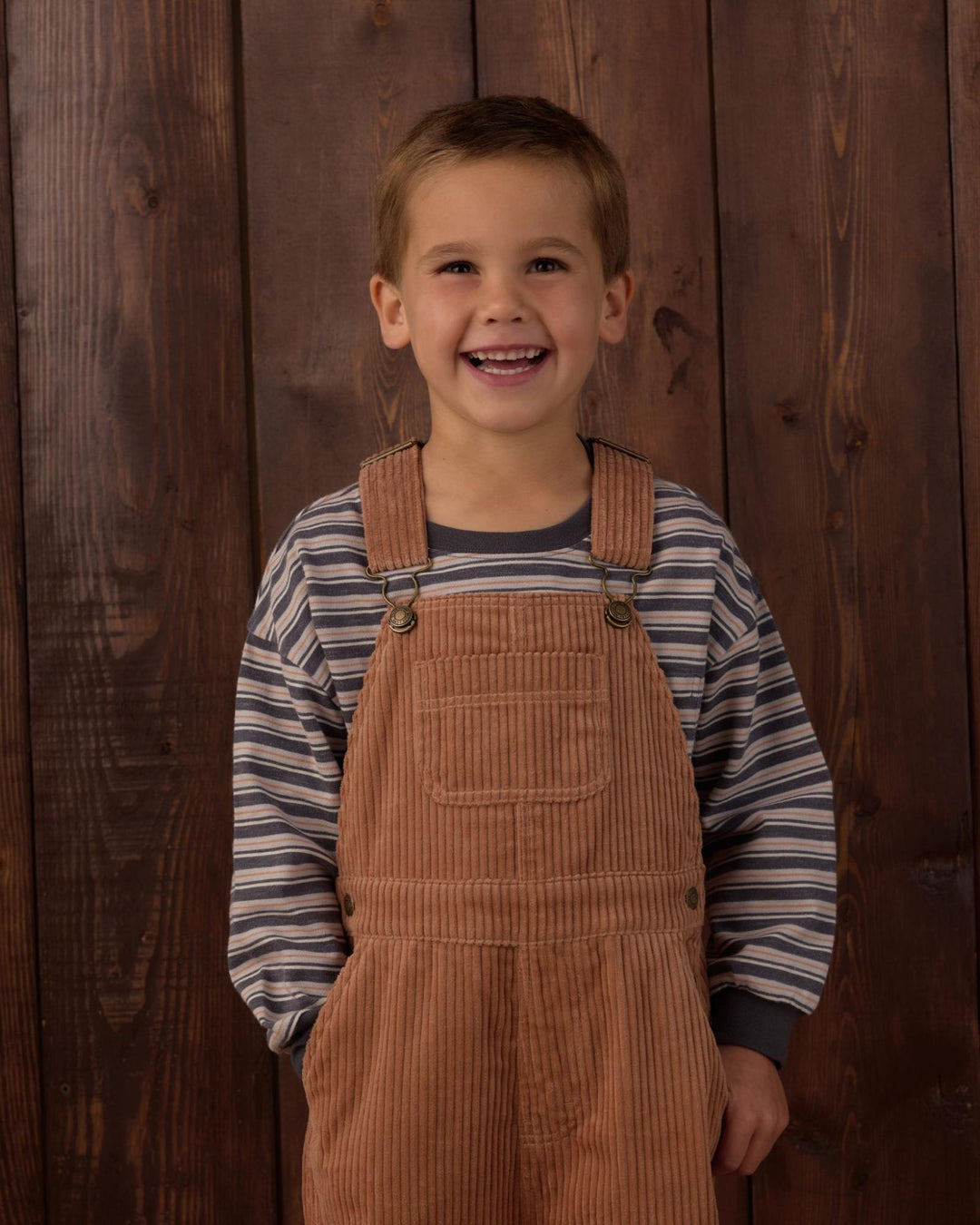 A young boy smiling in front of a dark wood plank background, wearing brown corduroy overalls over a navy and white striped long-sleeve shirt.