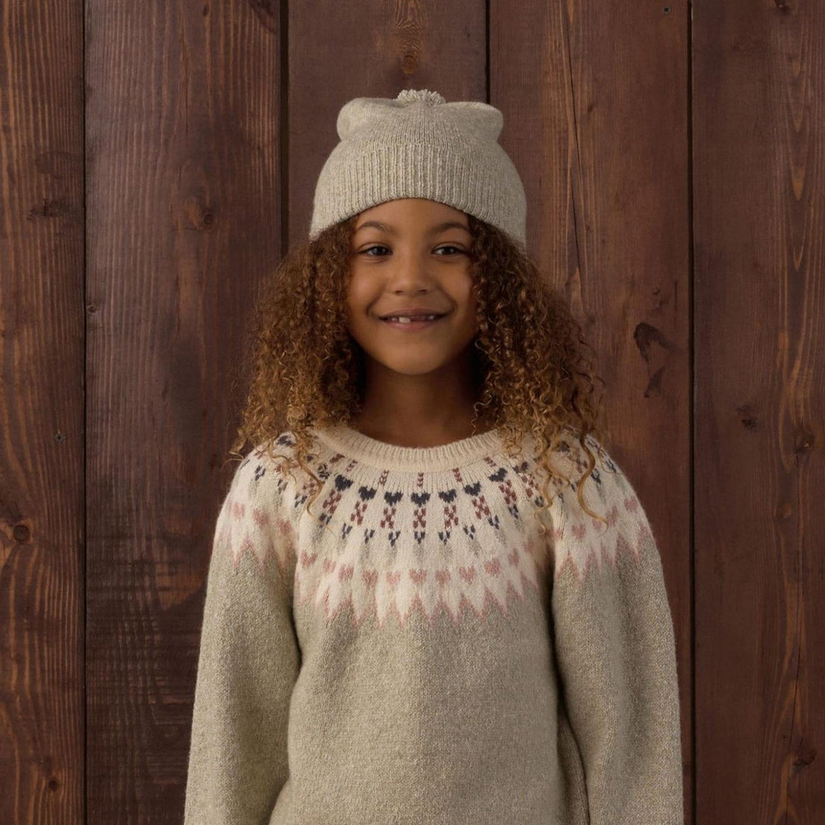 A young girl with curly hair stands against a dark wood plank wall wearing a cream knit sweater with a geometric Fair Isle pattern, a matching cream knit beanie hat, and a blue denim skirt.