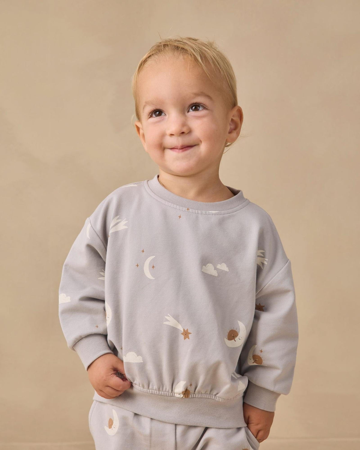 A smiling toddler with blonde hair wearing a light gray long-sleeve sweatshirt and matching pants with a celestial print of white clouds, moons, and stars, set against a plain beige background.