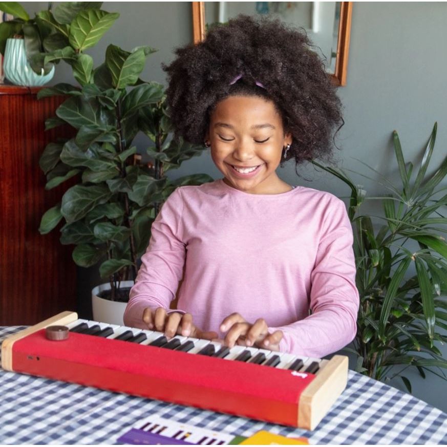 A young girl with an afro hairstyle is smiling and playing a small red toy keyboard with wooden keys on a checkered tablecloth, with potted plants in the background.