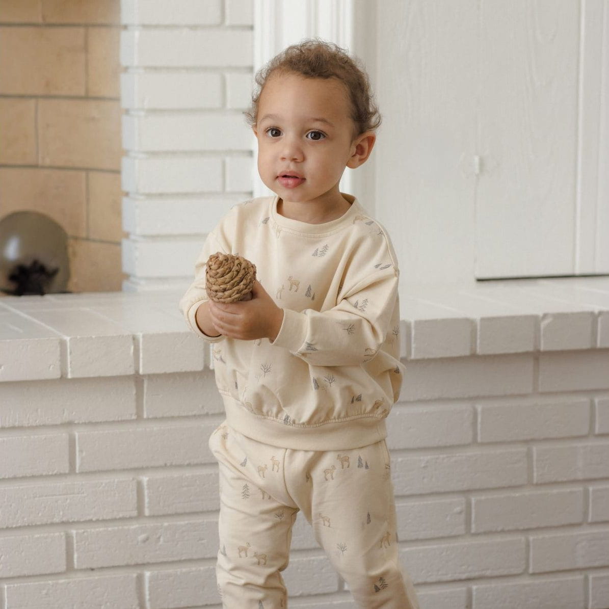 A toddler stands barefoot indoors in front of a white brick fireplace, wearing a cream-colored long-sleeve pajama set with a subtle allover animal print. The child is holding a small pinecone.