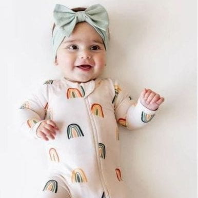 A baby lying on their back wearing a cream-colored footed sleeper with a rainbow pattern and a mint green bow headband, set against a plain white background.